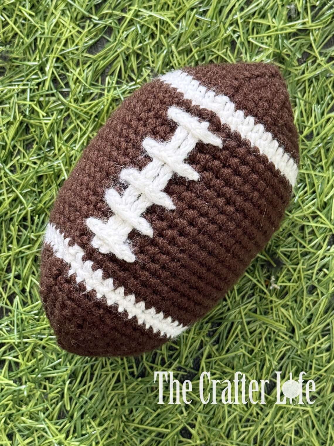 A textured brown crochet plush football with stitched white laces resting on bright green artificial grass turf.