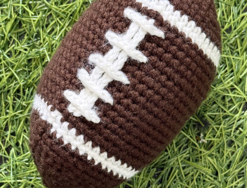 A textured brown crochet plush football with stitched white laces resting on bright green artificial grass turf.