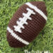 A textured brown crochet plush football with stitched white laces resting on bright green artificial grass turf.